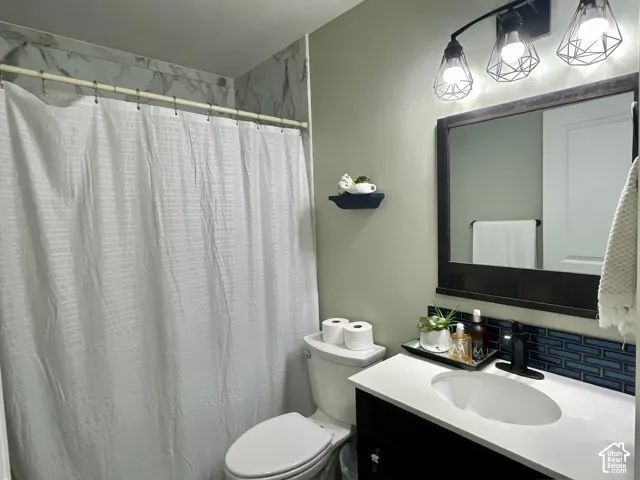 Bathroom featuring a vanity with a white countertop and oval under-mount sink, dark tile backsplash, three-light geometric wall fixture, and shower enclosure with decorative wall tile