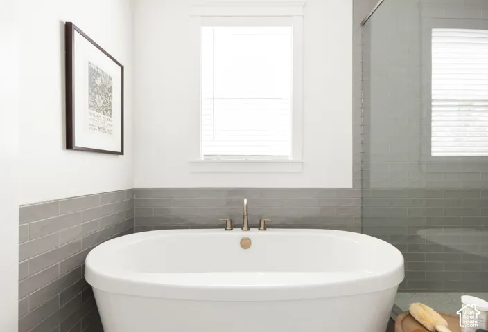Freestanding soaking tub with brushed brass fixtures, grey subway tile wainscoting, and an adjacent glass-enclosed shower