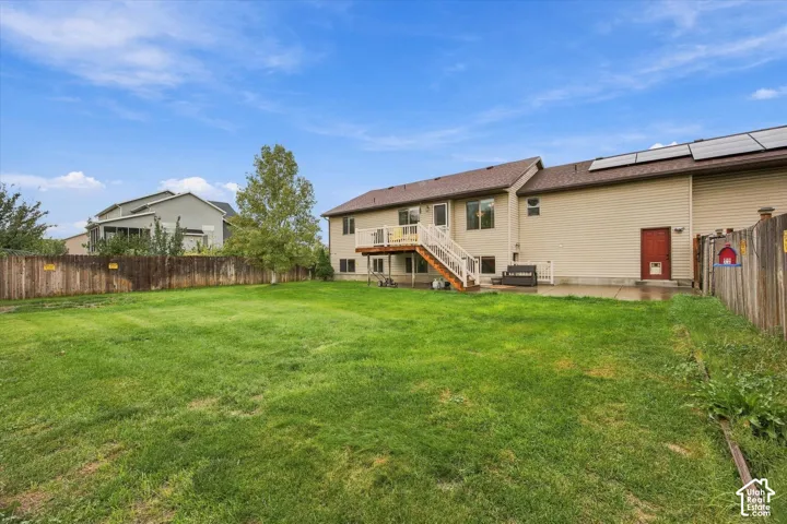 Expansive backyard featuring a manicured lawn, perimeter wood fencing, elevated wood deck with stairs, concrete patio, and roof-mounted solar panels