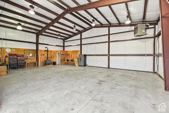 Spacious workshop featuring exposed steel beam construction, insulated wall panels, concrete flooring, overhead pendant lighting, and a solid-fuel stove