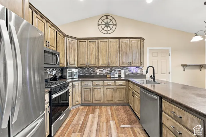 Kitchen featuring vaulted ceilings, wood-finish cabinetry, stainless steel appliances, multi-toned tile backsplash, and a peninsula with a matte black faucet