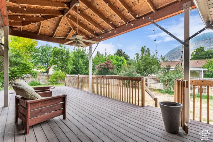 Covered wooden deck featuring exposed rafters, a ceiling fan, and perimeter wood balustrades, overlooking an expansive backyard with mature trees and mountain scenery