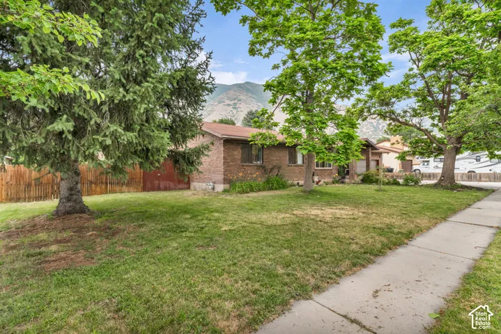Brick exterior residence featuring a front lawn, mature trees, wood fencing, and mountain scenery