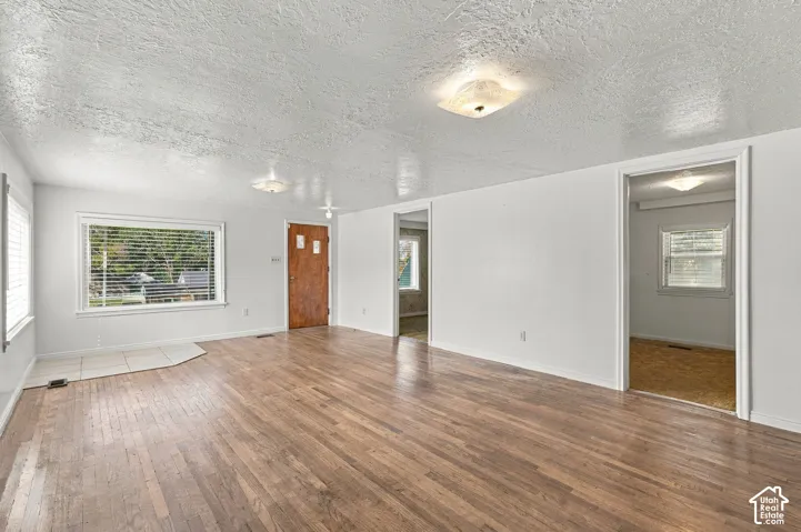 Wood-finish flooring, white painted walls, large window with blinds, front entry door, and ceiling-mounted light fixtures