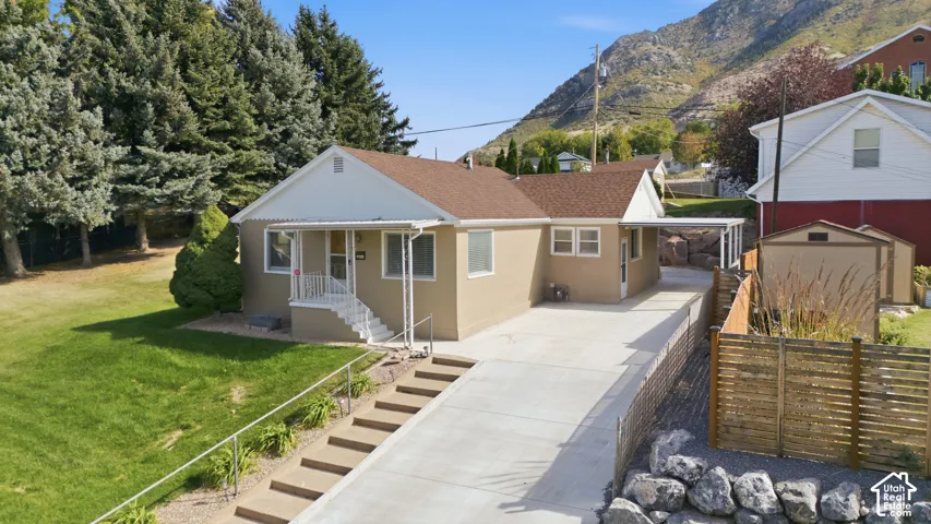 Stucco exterior with a brown shingle roof, covered front porch, concrete driveway, concrete front steps, and a landscaped front yard with mature evergreens