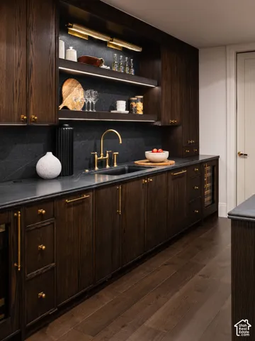 Wood-finish flooring, dark wood cabinetry, integrated beverage refrigerator, under-cabinet lighting, and a gold-finish faucet