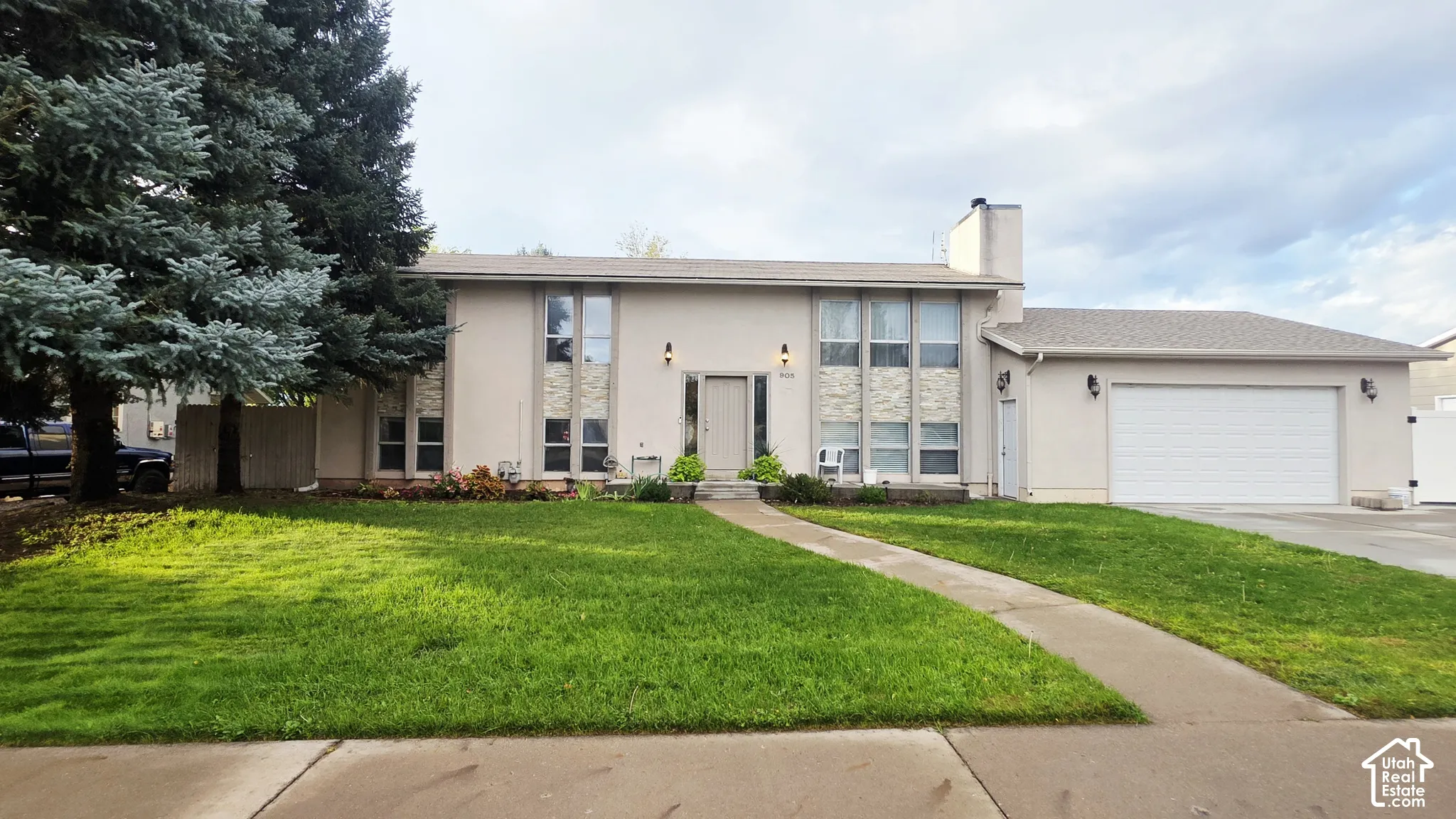 Multi-level exterior featuring light-toned stucco siding, stone veneer accents, multi-pane vertical windows, an attached two-car garage, and concrete walkway