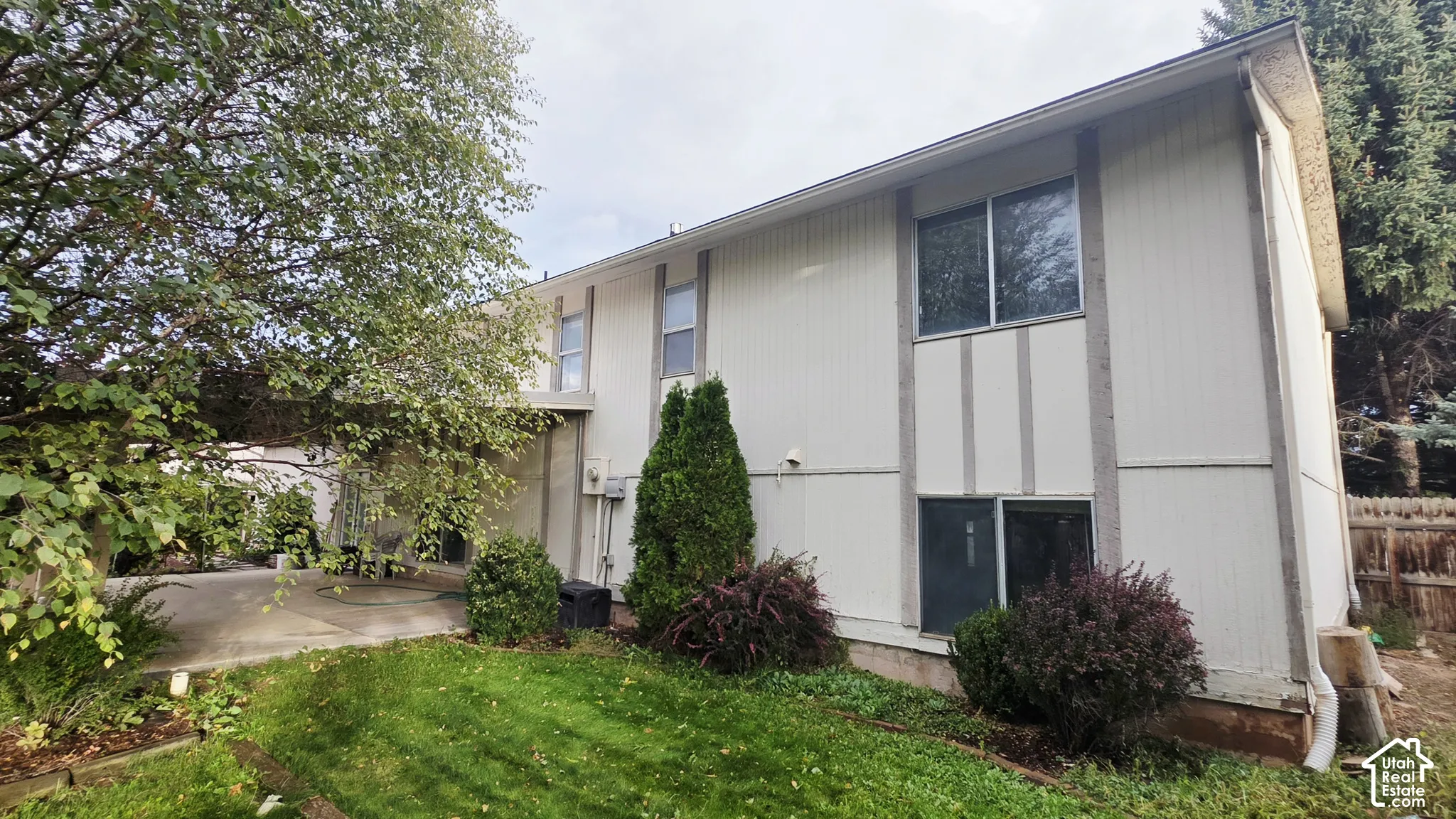 Two-story vertical siding exterior featuring a concrete patio, mature deciduous trees, evergreen shrubbery, and wood-panel fencing