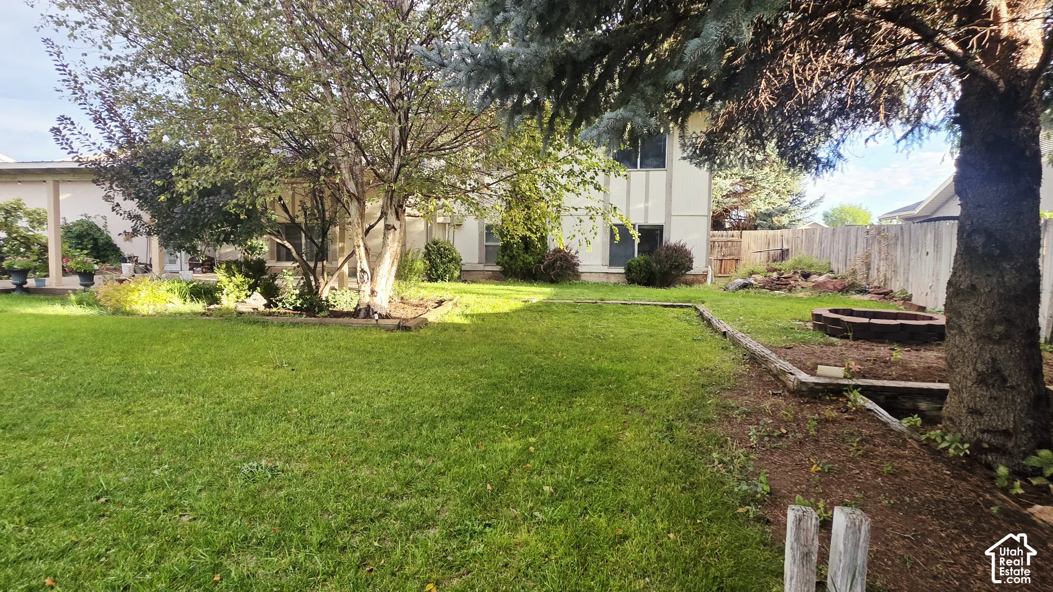 Backyard featuring a manicured lawn, established mature trees, tiered timber landscaping borders, and a circular stone fire pit