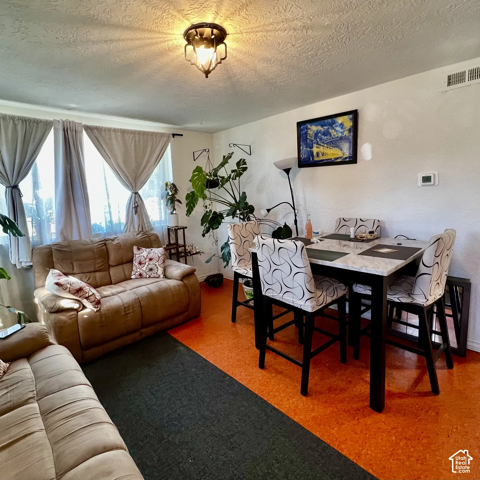 Bright living and dining area featuring a cognac leather sofa, black dining table with upholstered chairs, textured area rugs in complementary tones, large windows with layered curtains allowing natural light, and a decorative ceiling fixture