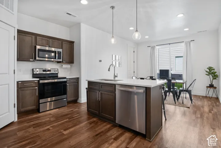 Kitchen featuring dark-toned cabinetry, stainless steel appliances, quartz countertops, a central island with an integrated sink, and wood-finish flooring