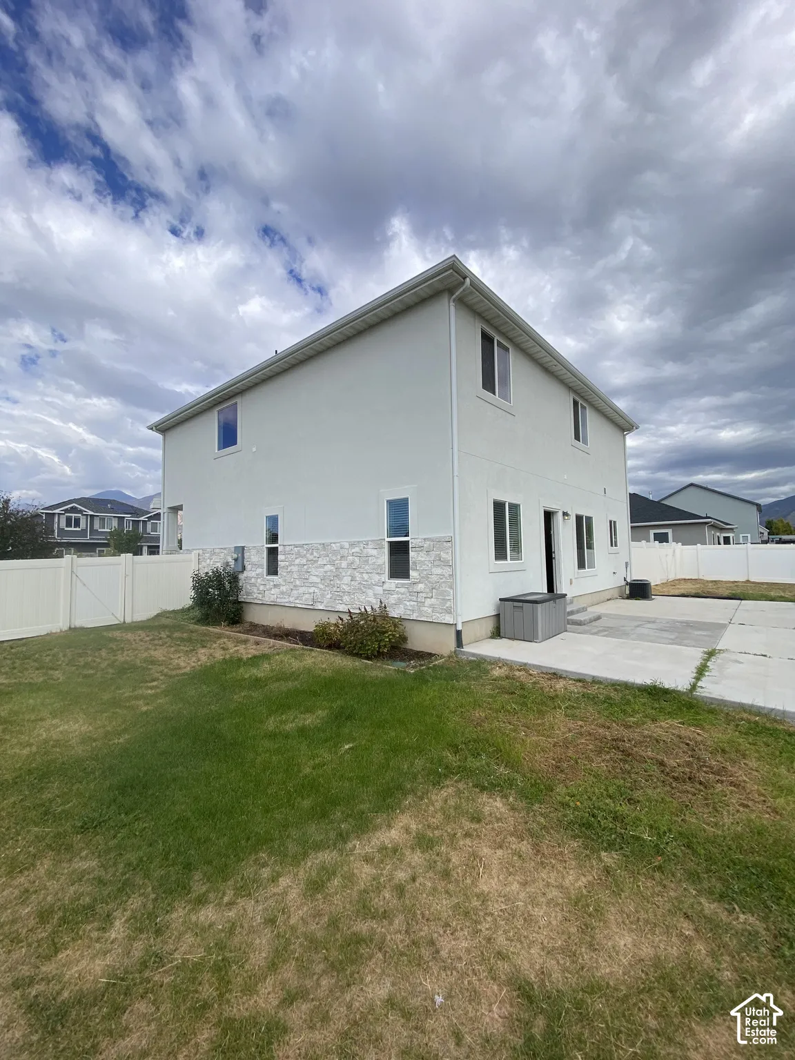 Exterior featuring light-toned stucco siding, natural stone veneer base, rear concrete patio, and white vinyl privacy fencing