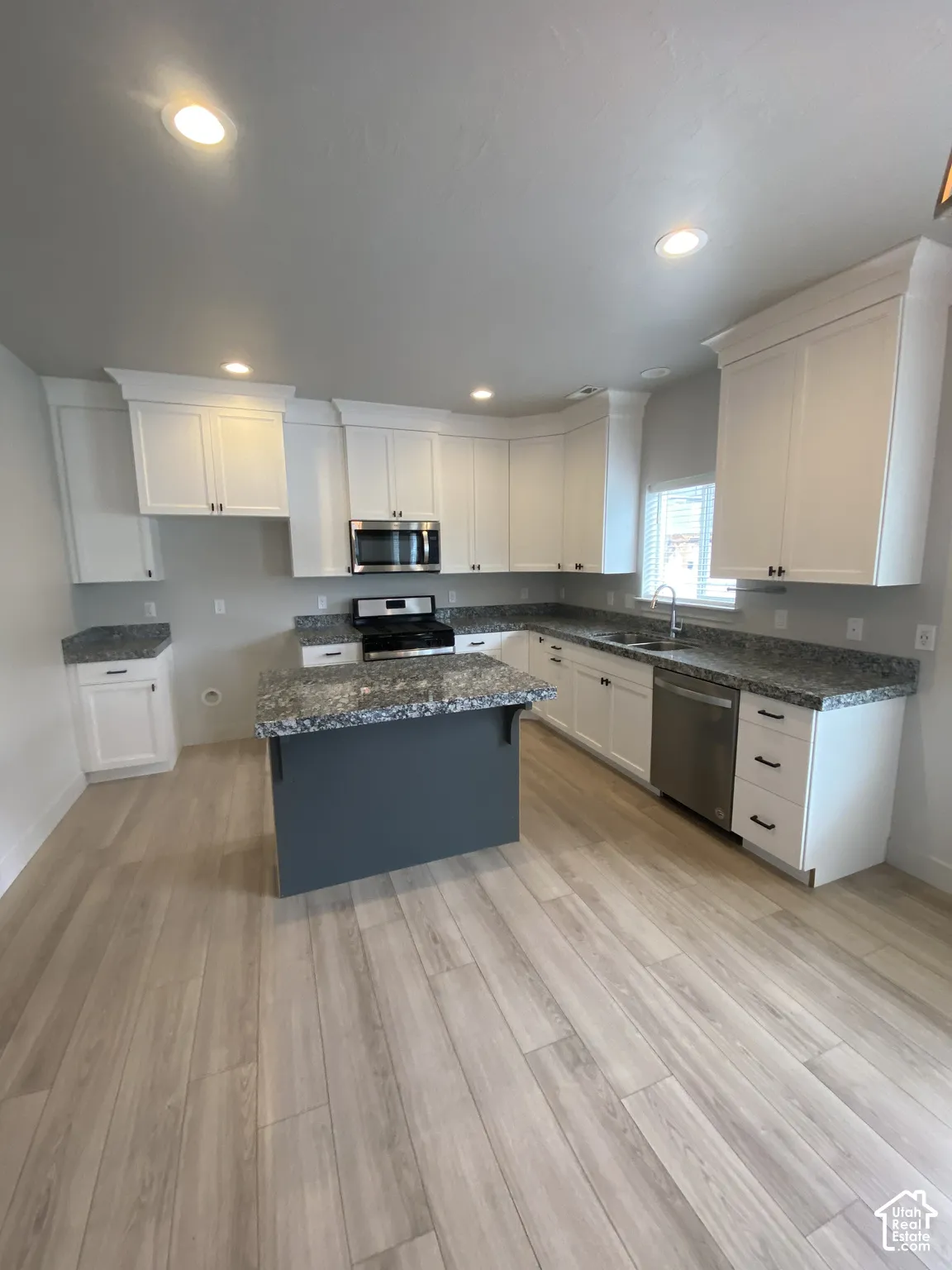 Kitchen featuring white shaker-style cabinetry, an island with a contrasting dark base, stone-patterned countertops, stainless steel appliances, and wood-finish flooring