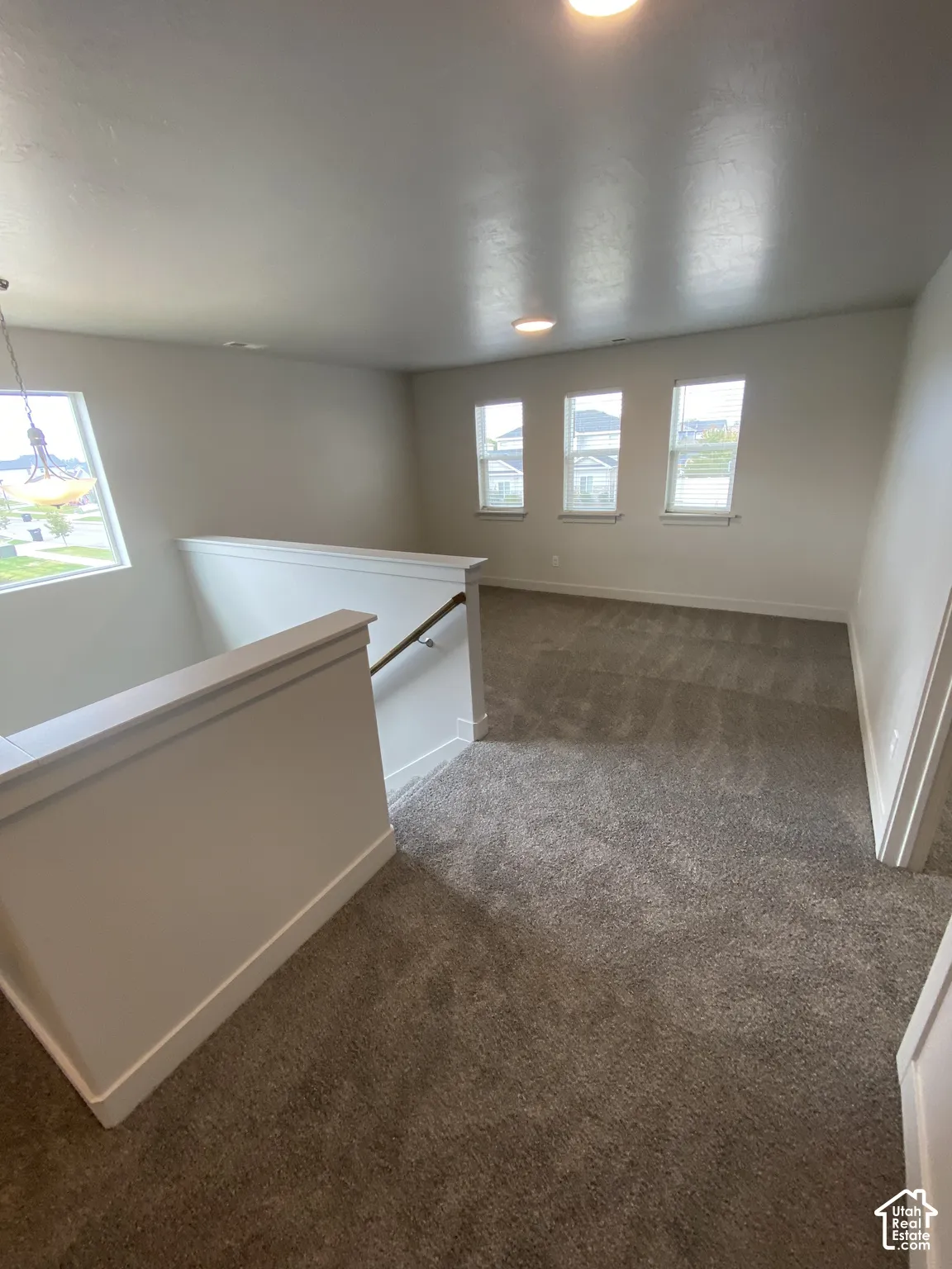 Open loft area featuring neutral wall-to-wall carpeting, multiple windows providing natural light, and a staircase with a wooden handrail