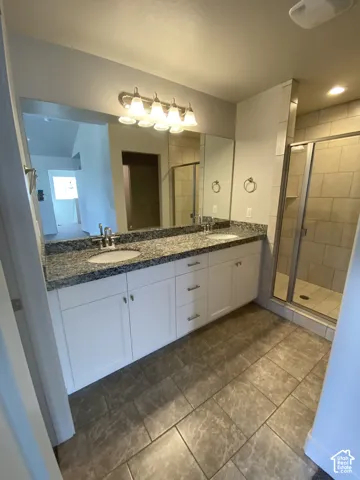 Dual-sink vanity with white shaker-style cabinetry, stone-finish countertops, tile flooring, and a walk-in shower with tiled surround
