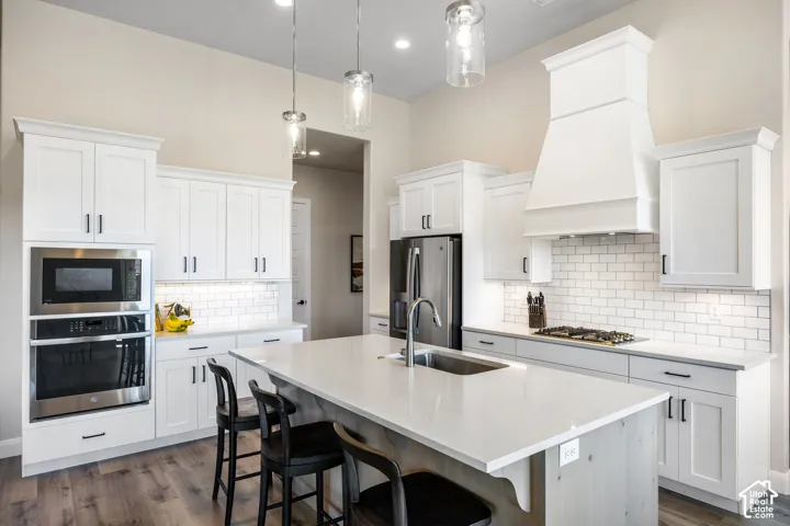 Kitchen featuring white shaker-style cabinetry, a central island with a stone countertop and under-mount stainless steel sink, subway tile backsplash, custom range hood, and stainless steel built-in double wall ovens