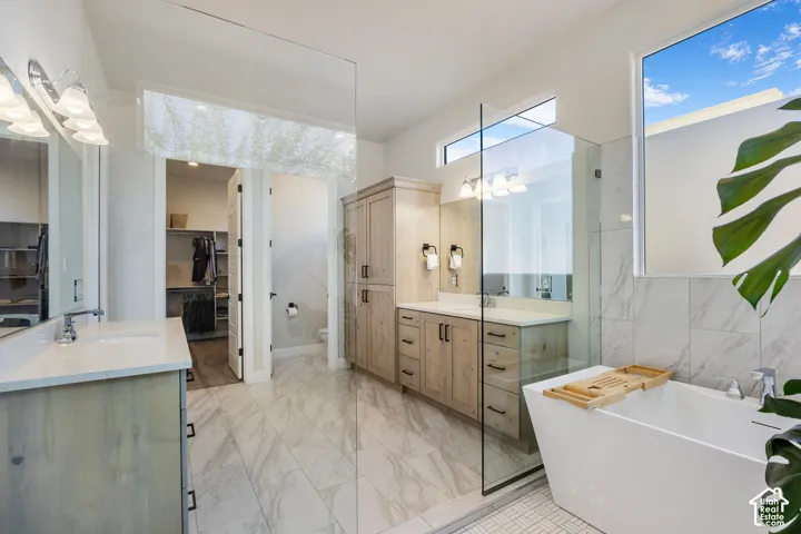 Bathroom featuring dual wood-finish vanities with light-toned countertops, a glass-enclosed walk-in shower, a freestanding soaking tub, and large-format tile flooring