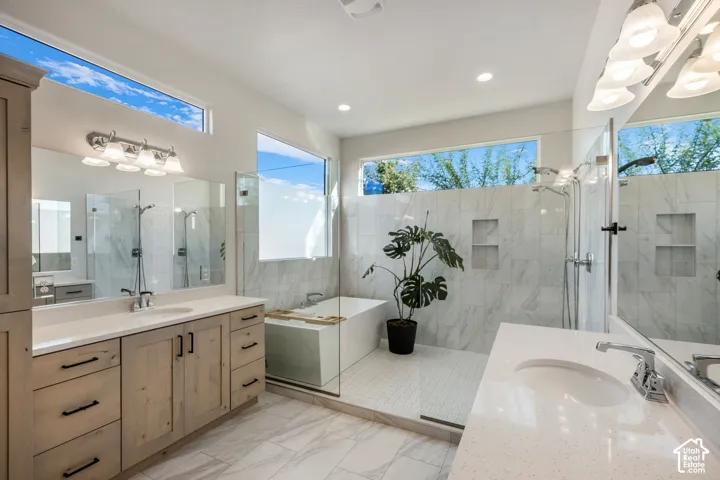 Dual-vanity bathroom featuring light-finish cabinetry with matte black hardware, quartz countertops, a glass-enclosed wet room with a soaking tub and rainfall shower, floor-to-ceiling stone-style tile, and clerestory windows