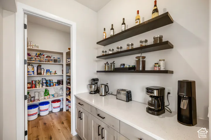 Walk-in pantry featuring custom built-in shelving, complemented by an auxiliary food preparation area with quartz countertops, wood-finish cabinetry, matte black hardware, and wall-mounted floating shelves