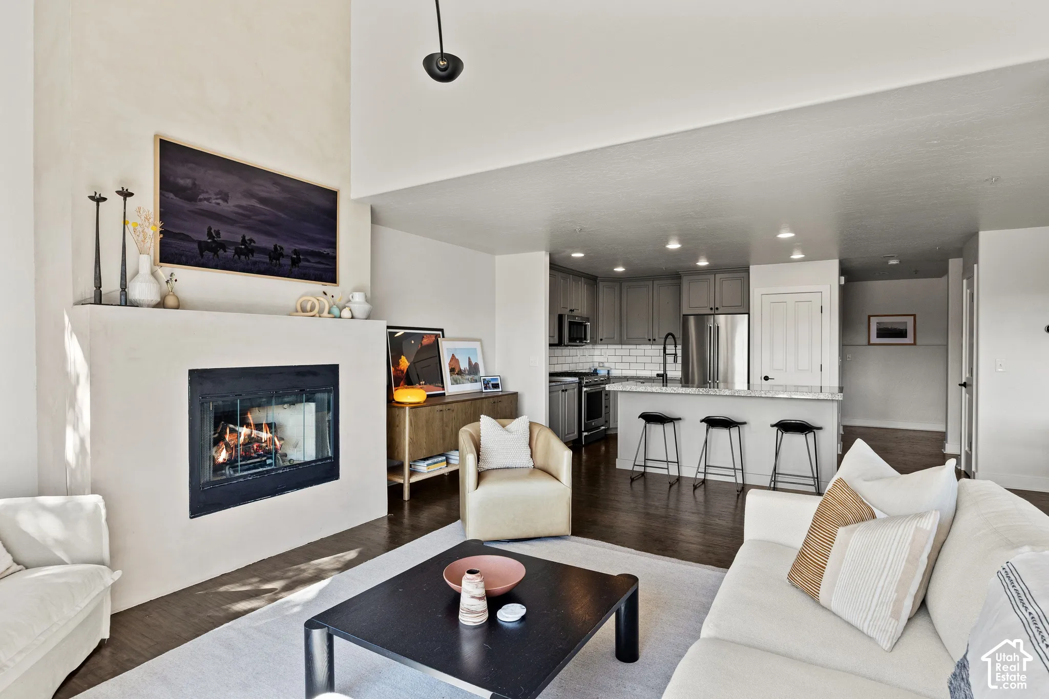 Living area featuring a fireplace with a stone surround, wood-finish flooring, and a kitchen with gray cabinetry, stainless steel appliances, and a breakfast bar with seating