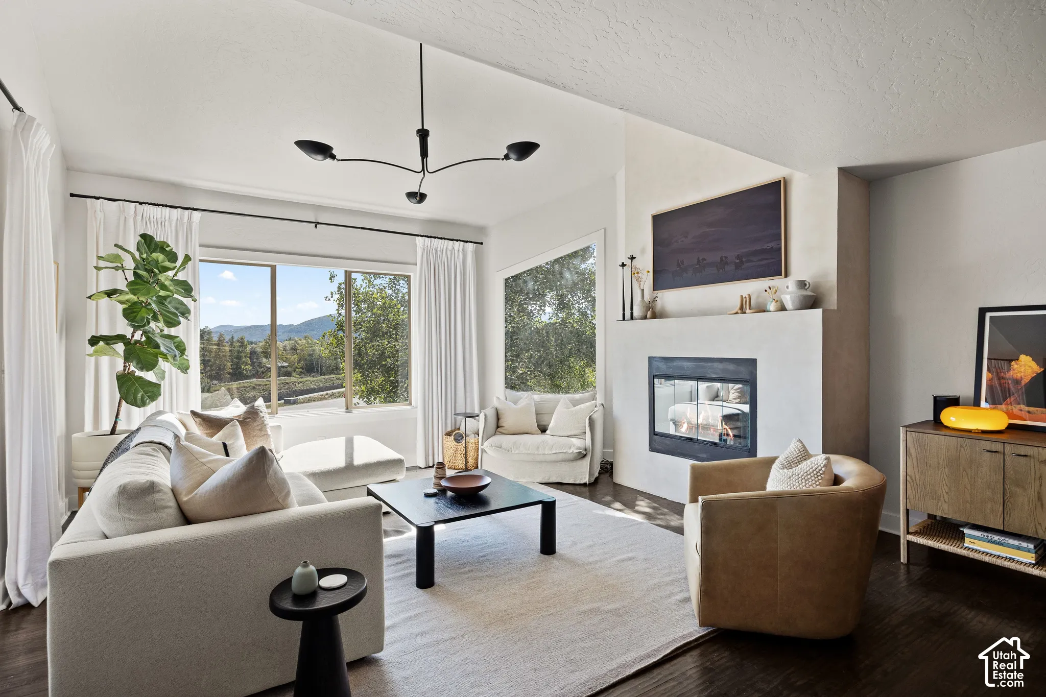 Wood-finish flooring, a contemporary fireplace with a dark-framed insert, and large windows with surrounding scenery