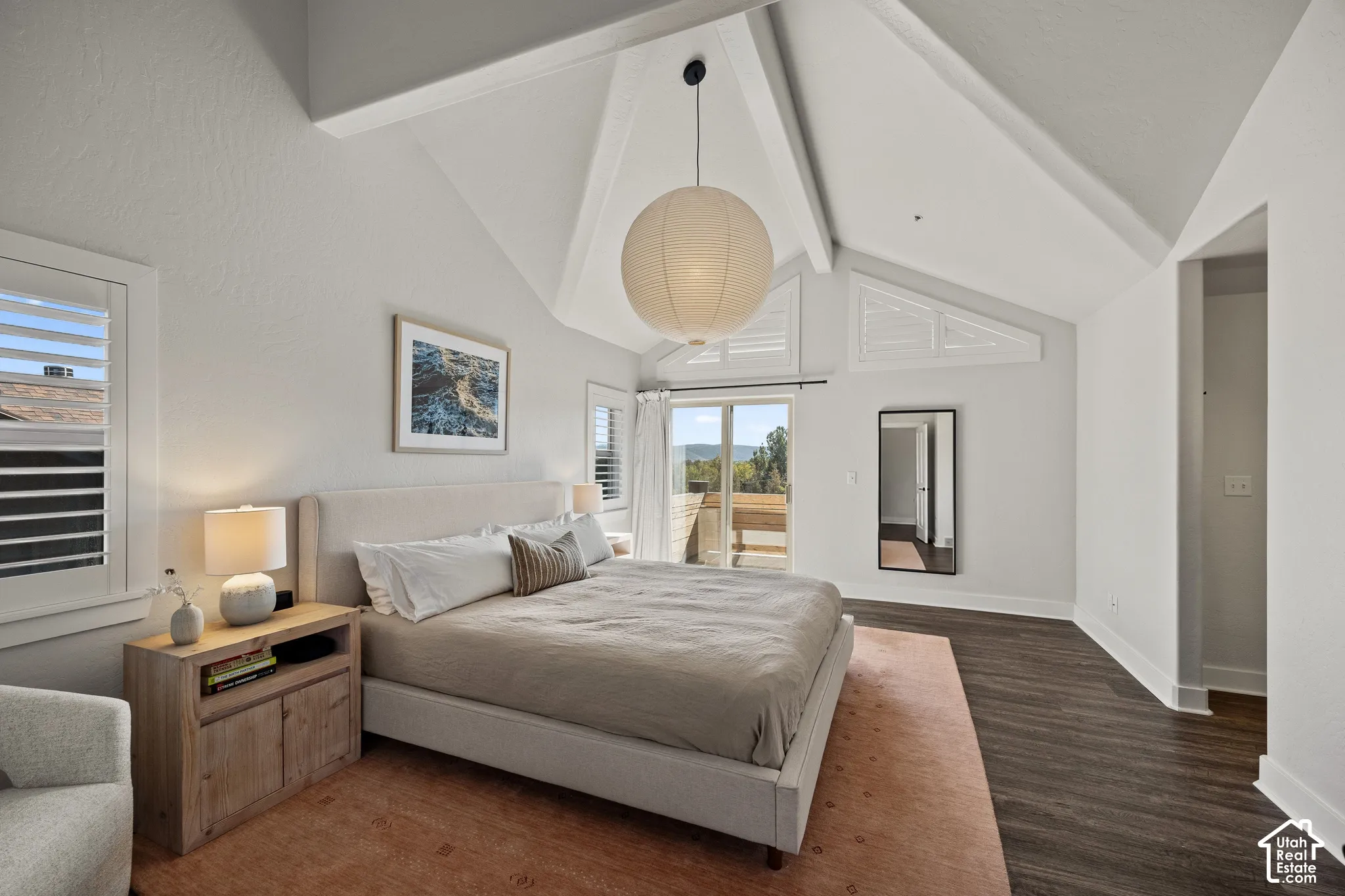 Vaulted ceiling with exposed beams, dark wood-finish flooring, white plantation shutters, and sliding glass door to exterior