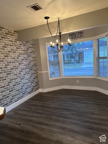 Exposed brick accent wall, wood-finish flooring, and bay window featuring horizontal blinds, finished with a multi-arm metal chandelier