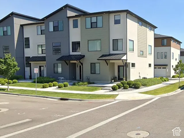 Multi-story exterior featuring a mix of stucco and horizontal siding, metal awnings over entryways, and concrete walkways surrounded by landscaped shrubbery