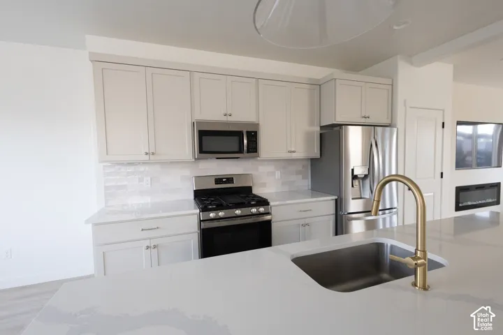 Kitchen featuring light-toned cabinetry, light-toned countertops, tile backsplash, stainless steel appliances, and a gold-tone faucet with an undermount sink