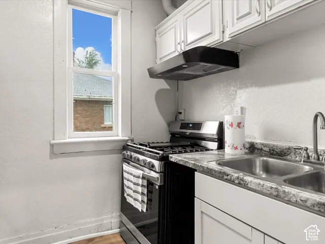 Kitchen featuring white shaker-style cabinetry, a stainless steel gas range, a black over-range vent hood, patterned laminate countertops, and a dual-basin stainless steel sink