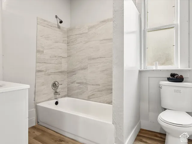 Bathroom featuring a tub with a light-toned, textured stone-finish surround, wood-finish flooring, chrome plumbing fixtures, and a dual-pane window