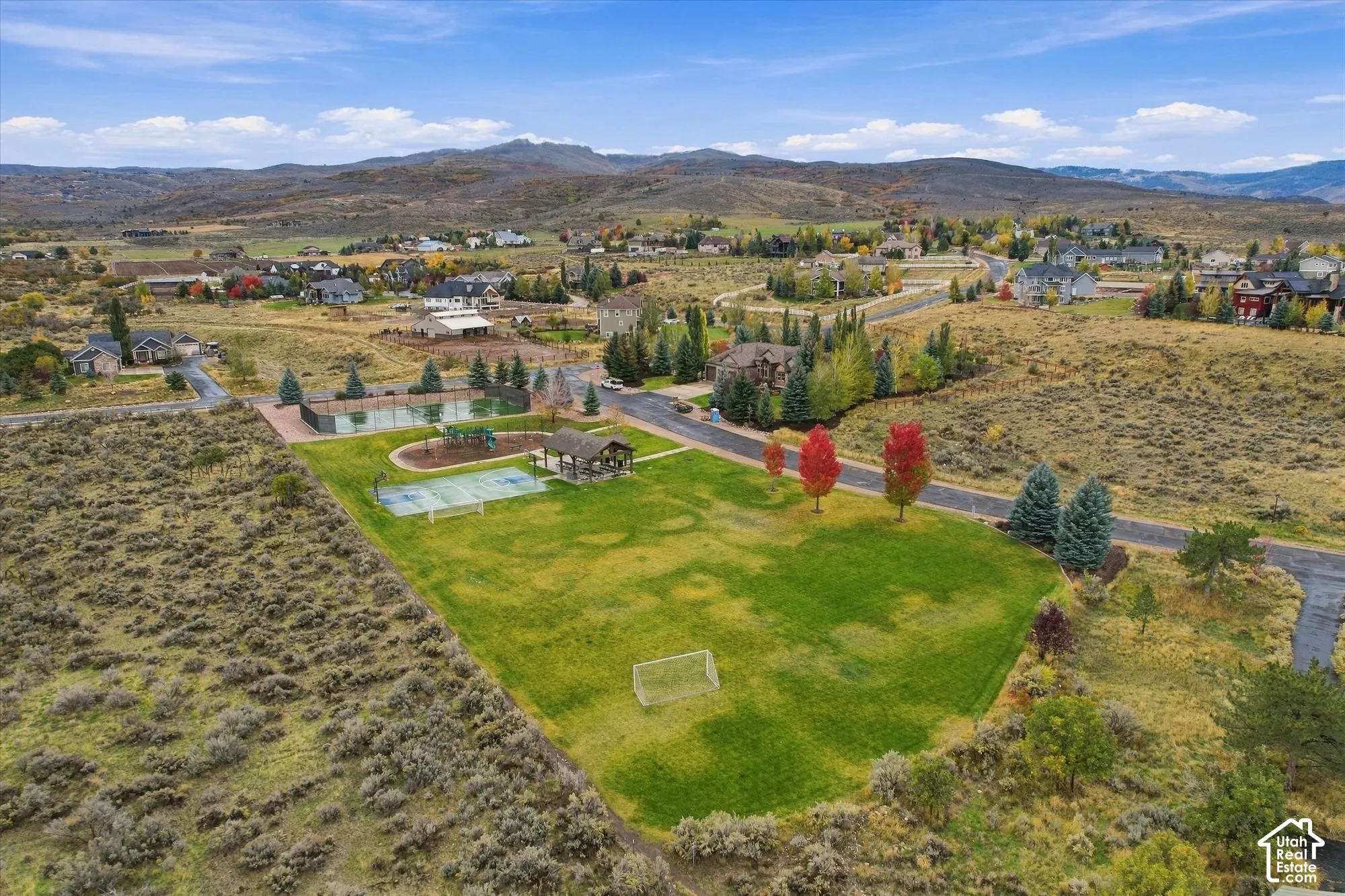 Expansive community grounds featuring a manicured lawn, multi-purpose sports court, paved tennis court, shaded gazebo, and a mountainous landscape backdrop