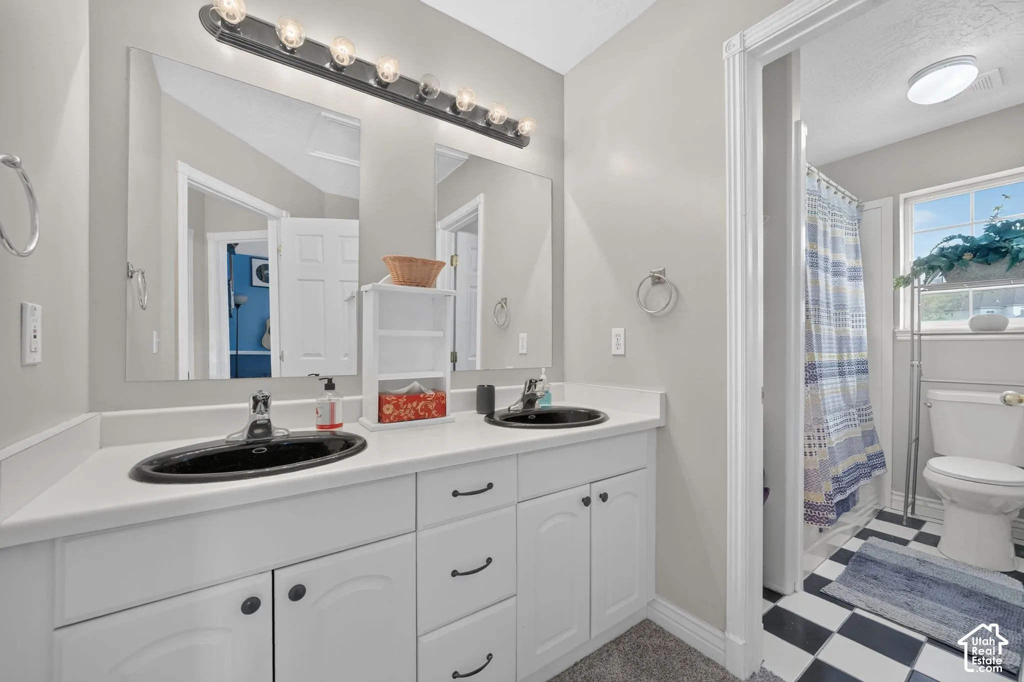 White dual-sink vanity with matte black hardware, integrated rectangular mirrors, expansive lighting fixture, and checkerboard floor tile