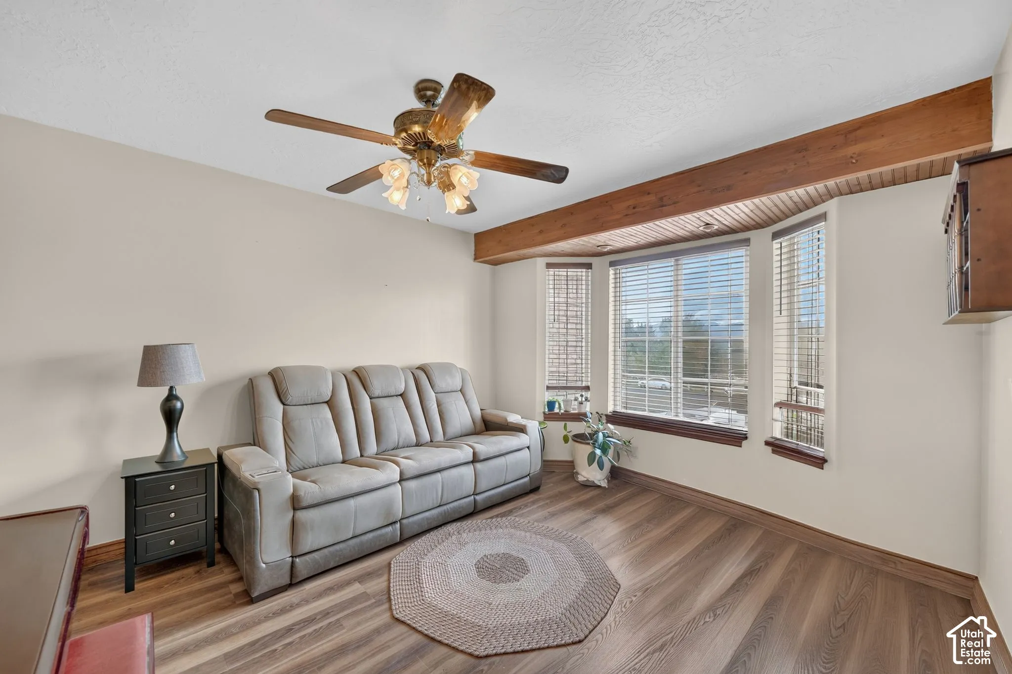 Wood-finish flooring, bay window with integrated blinds, exposed wood ceiling beam, wood-paneled ceiling accent, and ceiling fan with integrated lighting