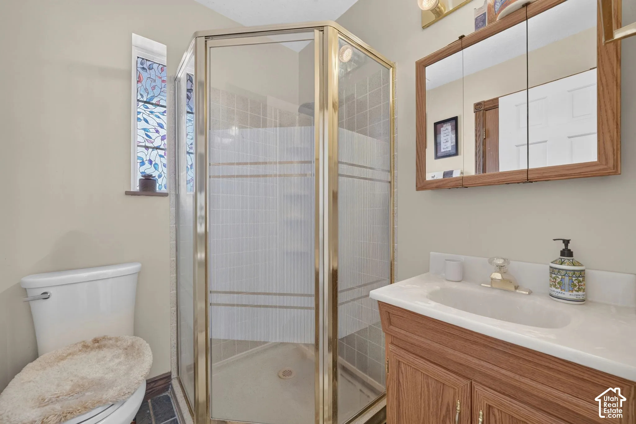 Corner shower stall with gold-tone frame and tile surround, wood-finish vanity with integrated sink and countertop, and decorative stained-glass window accent