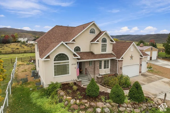Multi-story residence featuring a stone and stucco facade, arched window detailing, multi-gabled roofline, tiered rock retaining wall, and an attached garage with a detached secondary structure