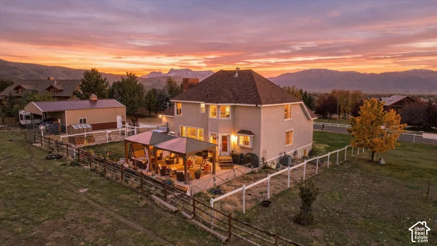 Multi-level stucco exterior featuring a detached workshop with carport, stone-paved patio with metal pergola, split-rail perimeter fencing, and expansive grounds set against a mountain backdrop