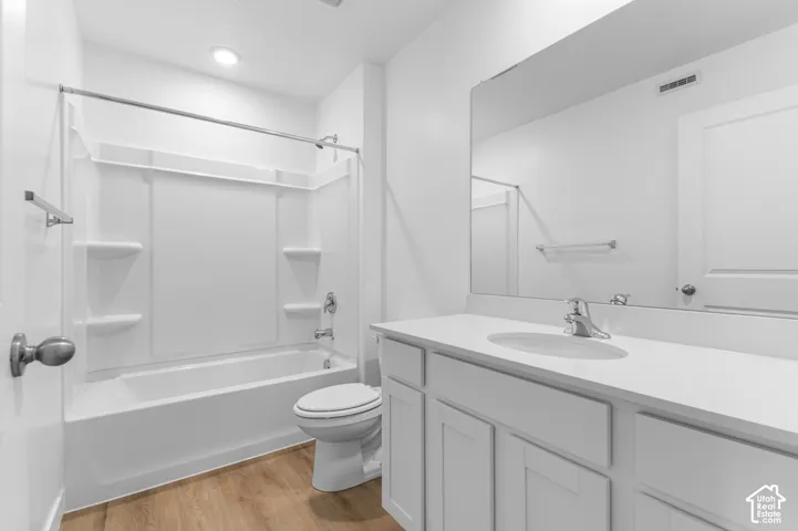 White vanity with a solid-surface countertop and integrated sink, large wall-mounted mirror, white shaker-style cabinetry, bathtub-shower combination with integrated shelving, and wood-finish flooring