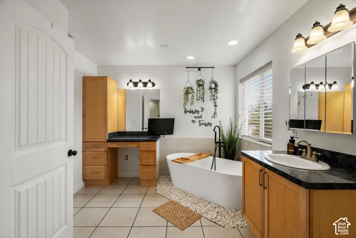 Freestanding soaking tub with floor-mounted faucet, stone-accented tub surround, built-in wooden vanity with dark stone-finish countertop, integrated tall storage cabinetry, and tiled flooring