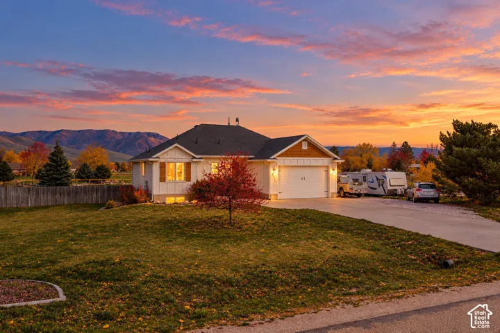 Single-story residence featuring a mixed-material facade of vertical siding and stone accents, concrete driveway, attached garage, and mountain backdrop and digitally altered sunset