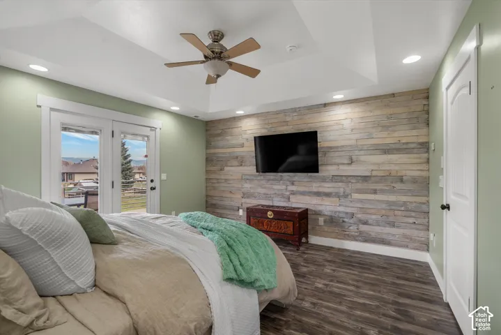 Tray ceiling featuring a central lighted ceiling fan, wood-plank accent wall, wood-finish flooring, and double glass-paned doors