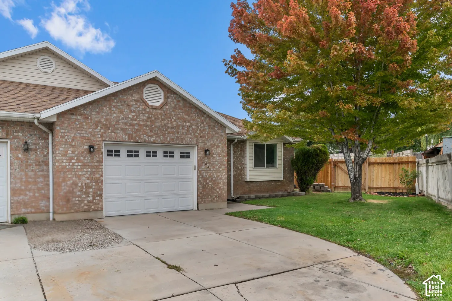 Brick exterior featuring an attached garage, concrete driveway, and a fenced lawn with mature tree