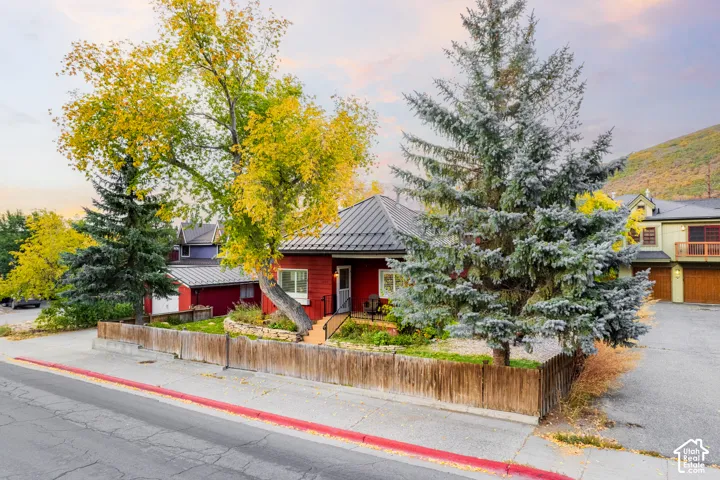 Red exterior with black metal roof, front yard with mature trees, wooden fence, and paved street access