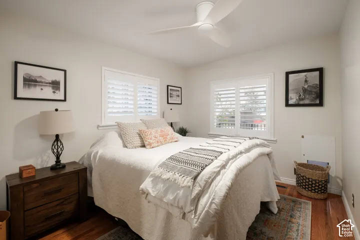 Hardwood flooring, white wall paint, two windows with plantation shutters, ceiling fan, and built-in wall outlets