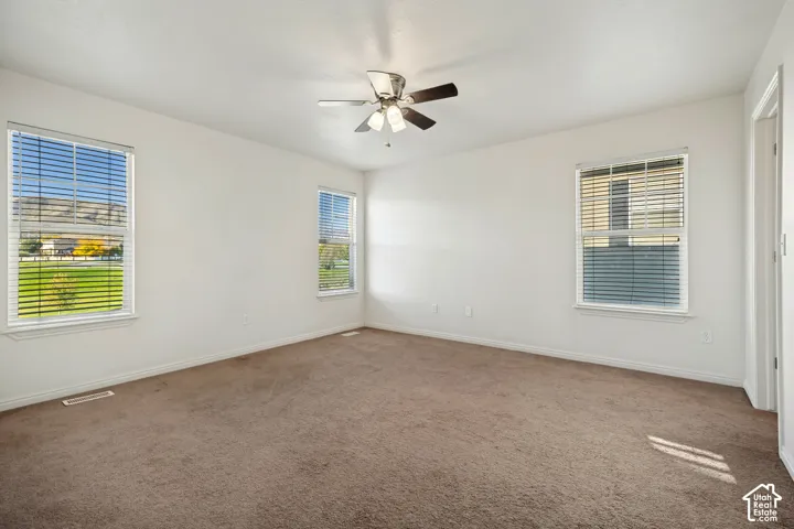 Neutral-toned carpeting, three windows with horizontal blinds, and a central ceiling fan with multi-light fixture