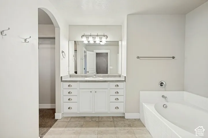 Bathroom featuring a white vanity with integrated cabinetry, an expansive wall-mounted mirror, a tiled deck-mount soaking tub, and tile flooring