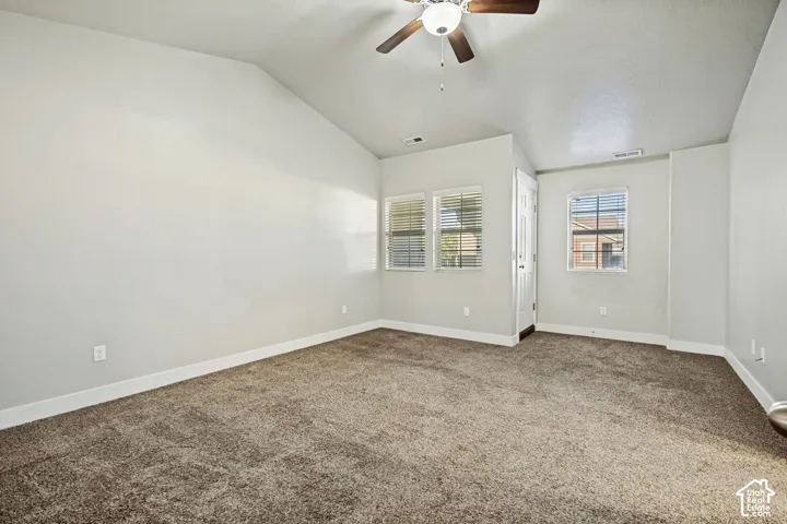 Vaulted ceiling featuring a central light fixture with a ceiling fan, neutral carpeting, and three windows