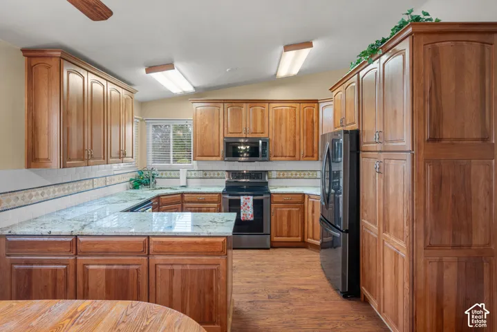 Kitchen featuring stained wood cabinetry, stone countertops, stainless steel appliances, decorative tile backsplash, and wood-finish flooring