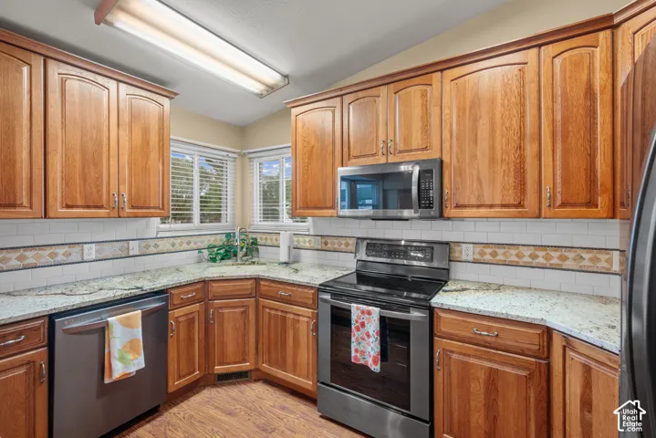 Kitchen featuring wood cabinetry, stone countertops, stainless steel appliances, subway tile backsplash with decorative inset, and wood-finish flooring