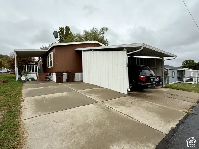 Exterior featuring a brown metal-sided facade, concrete driveway, covered carport with side privacy screening, and a raised entry platform with white railing